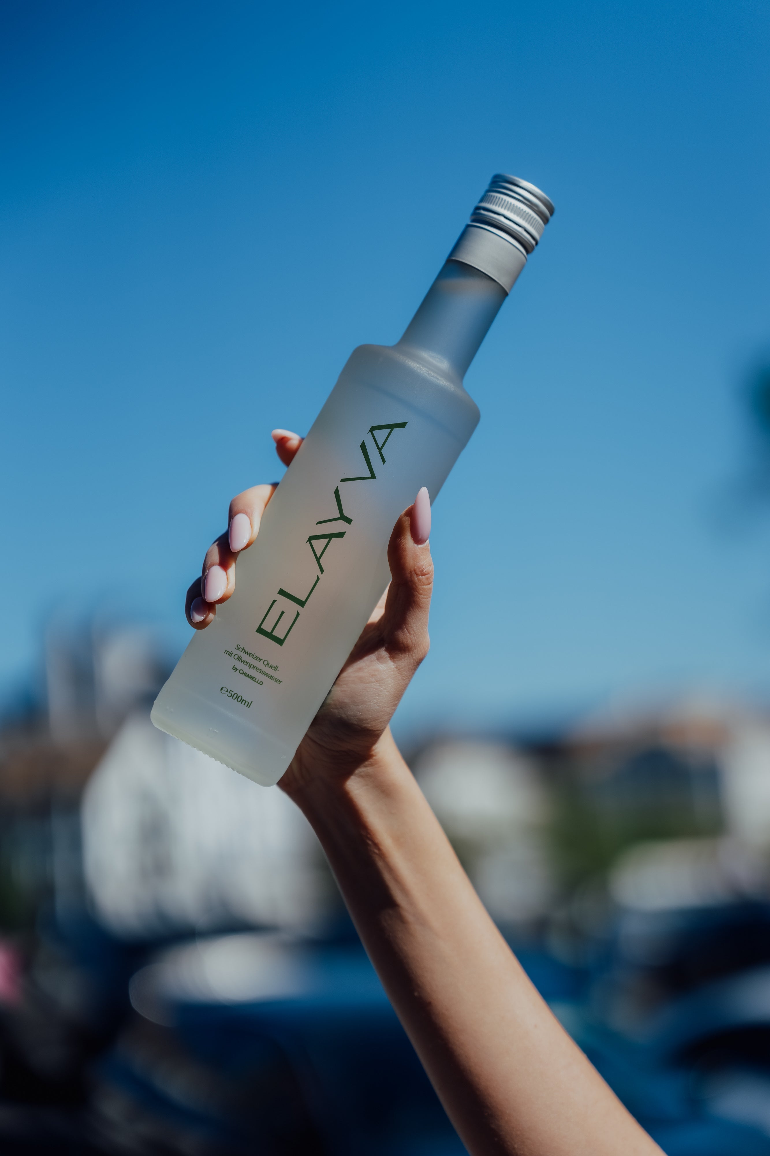Hand holding a frosted ELAYVA glass bottle against a soft blue outdoor backdrop